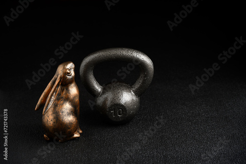 Fototapeta Naklejka Na Ścianę i Meble -  Holiday fitness and Happy Easter, gold metal bunny with a silver iron kettlebell on a black background looking up adoringly
