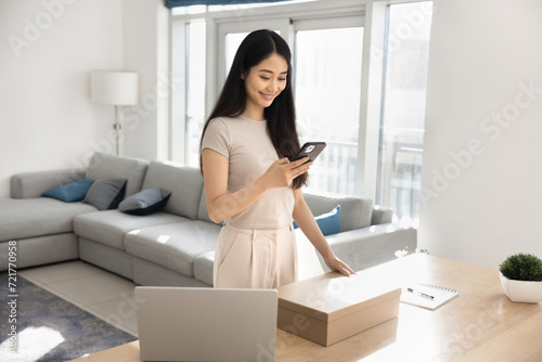 Cheerful young Chinese webstore customer girl using ecommerce app on mobile phone over cardboard box at home, making delivery order, texting shipping service online