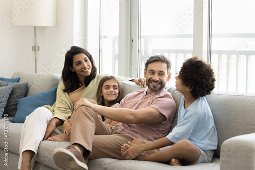 Happy mom and dad talking to preteen son and daughter on home couch, relaxing together in apartment, enjoying conversation, communication, discussion, smiling, laughing