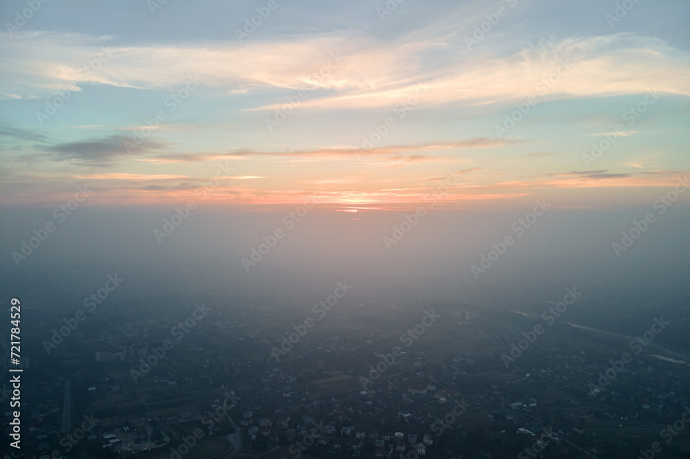 Aerial view from airplane window at high altitude of distant city covered with layer of thin misty smog and distant clouds in evening