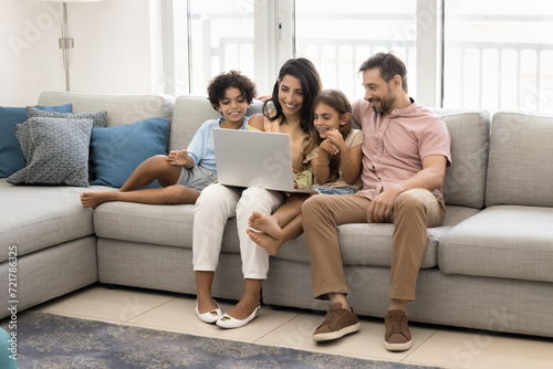 Cheerful sweet preteen kids and couple of parents enjoying leisure with laptop on spacious comfortable home couch, using smart technology, media service for watching movie