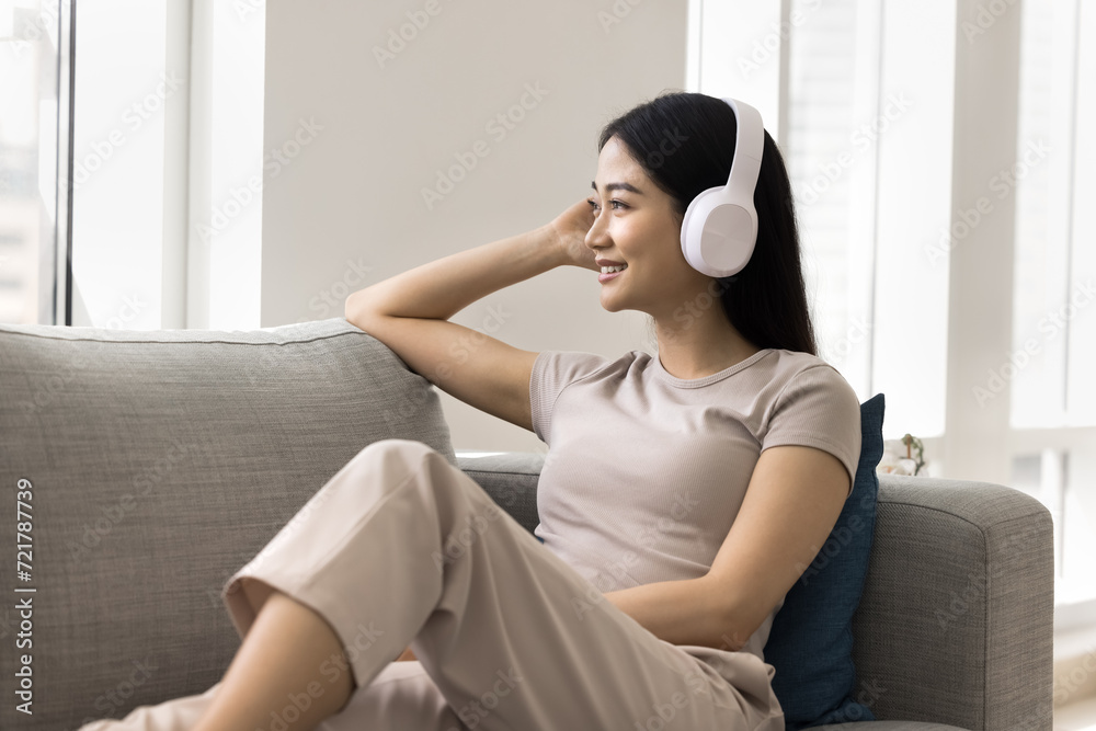 Positive relaxed young Asian music fan girl wearing wireless headphones, looking at window away, listening to songs from online playlist, musical Internet service, media application, relaxing at home Positive relaxed young Asian music fan girl wearing wireless headphones, looking at window away, listening to songs from online playlist, musical Internet service, media application, relaxing at home