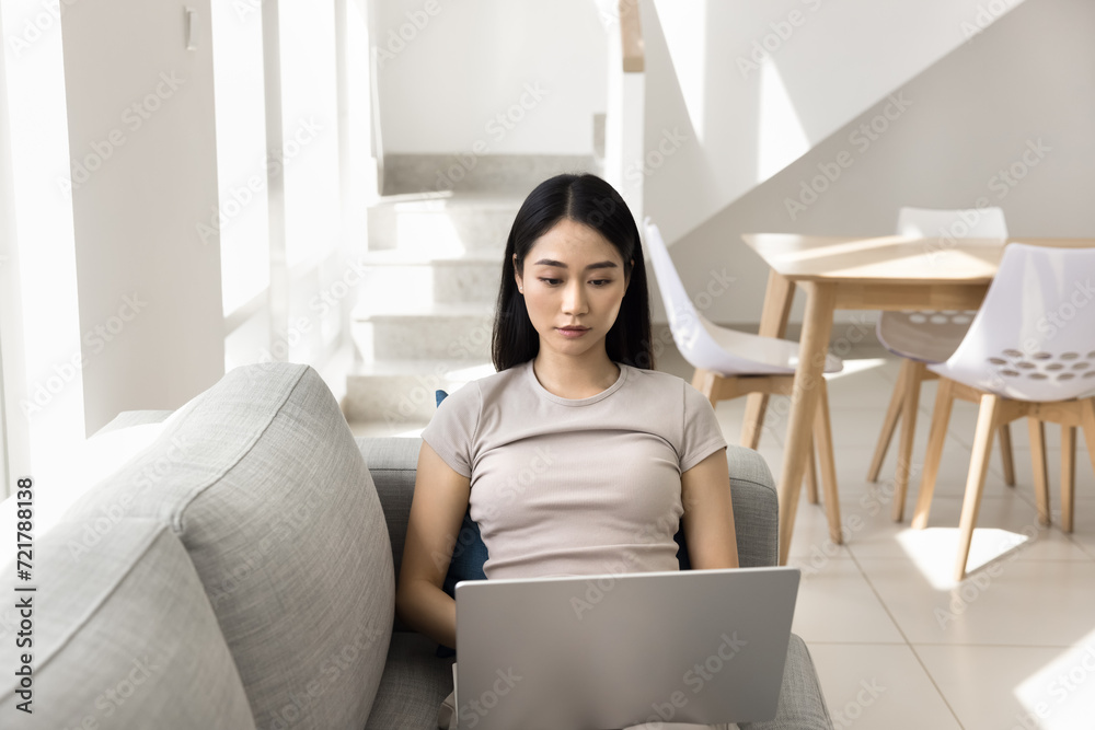 Focused young Asian freelance employee working at home, typing on laptop, resting on sofa, holding computer on lap, using online job, typing, reading, watching content on Internet Focused young Asian freelance employee working at home, typing on laptop, resting on sofa, holding computer on lap, using online job, typing, reading, watching content on Internet