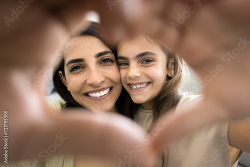 Happy little child girl and young mom looking at camera through finger heart, making hand frame, symbol of love, kindness, friendship, smiling, showing perfect white teeth, posing for family portrait © fizkes