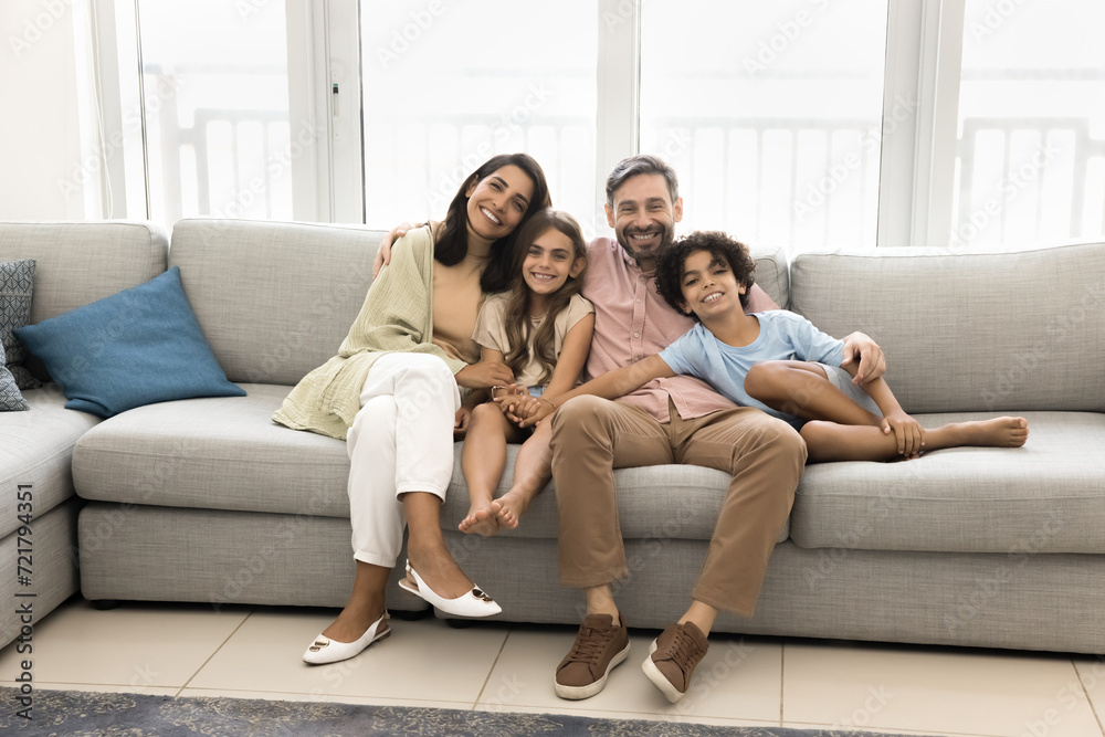Cheerful young parents and preteen children resting on home couch, looking at camera, smiling, enjoying being in comfortable new apartment, promoting mortgage, affordable property buying for family Cheerful young parents and preteen children resting on home couch, looking at camera, smiling, enjoying being in comfortable new apartment, promoting mortgage, affordable property buying for family