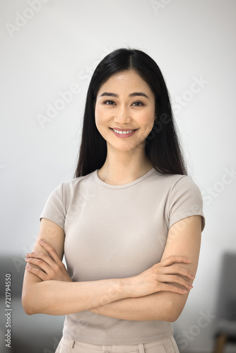 Cheerful confident beautiful Korean woman home vertical portrait. Happy young Asian female model in casual posing in apartment with hands folded, looking at camera with toothy smile