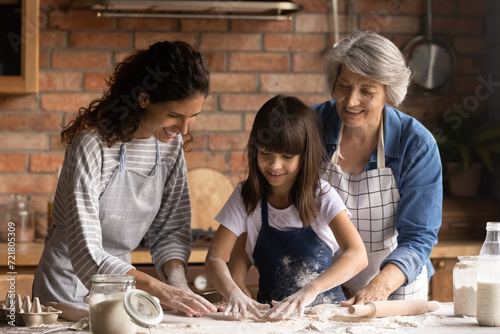 Hands in flour. Friendly intergenerational family of 3 diverse age females engaged in cooking short pastry biscuits. Young mom senior granny assist little girl stretch dough for pie on kitchen table