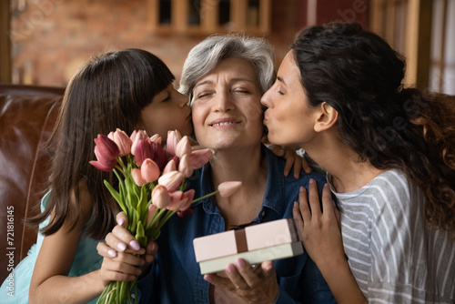 Happy Mothers Day. Affectionate young adult woman daughter and little kid girl granddaughter greeting excited mature latin lady mommy grandmother with birthday holiday kiss give flowers present gift photo