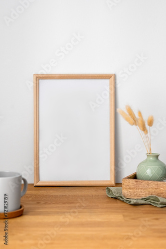 A frame with a blank canvas against a light wall and on a wooden tabletop with vase and cup of tea.
