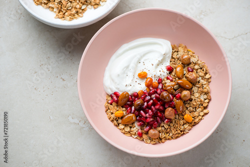 Roseate bowl with granola, yogurt, pomegranate and nuts, horizontal shot on a light-beige stone background, elevated view