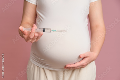Pregnant woman holding a syringe in her hand, studio pink background. Pregnancy and medical procedures concept