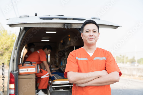Asian male ambulance staff member standing with crossed arms. He is wearing ambulance uniform of paramedics during paramedics giving injured person first aid background