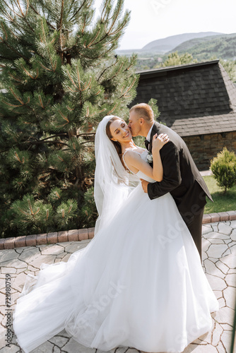 A handsome groom embraces his bride in a lush white dress and smiles in a beautiful outdoor setting. Under the open sky. High quality photo. A newlywed couple poses together on a sunny summer day.