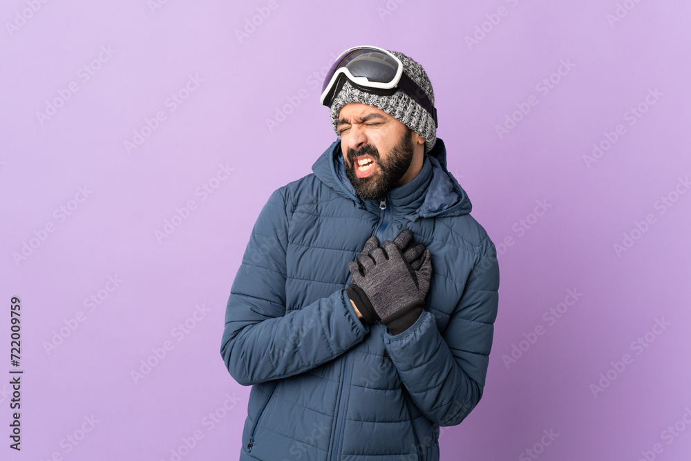 Skier man with snowboarding glasses over isolated purple background having a pain in the heart