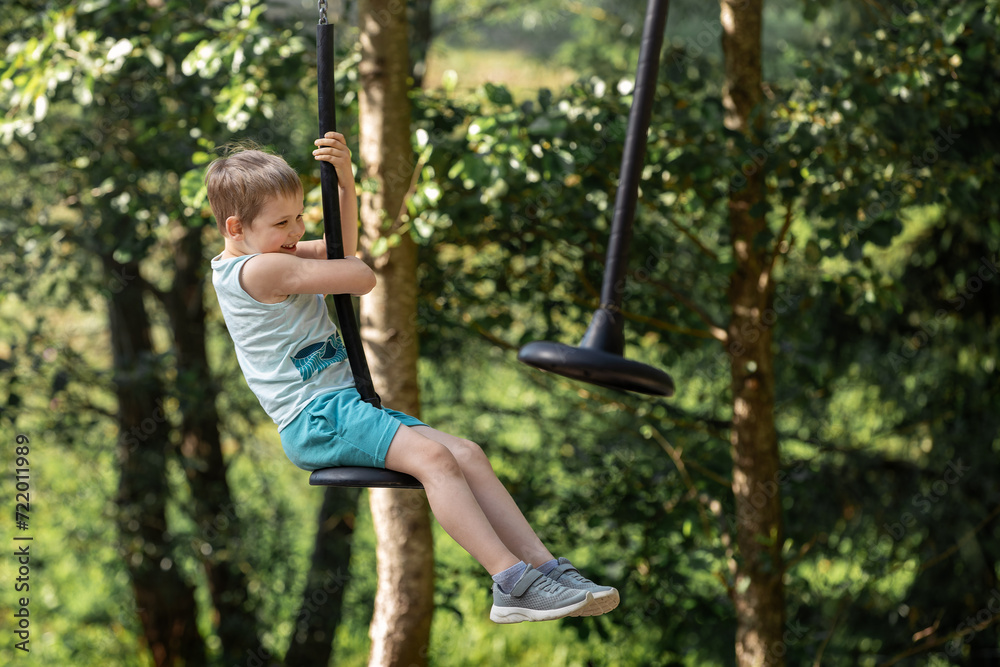 A little brave boy is rolling down a rope cable on a hanging seat between the trees of the summer forest.