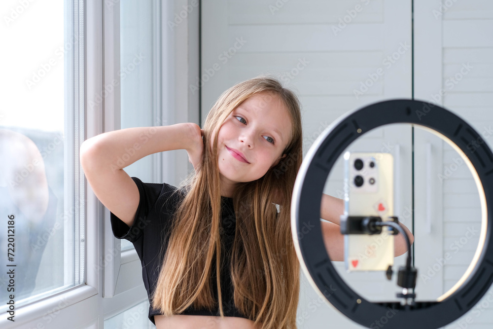 Beautiful teenage girl posing in front of a phone on a tripod. Child blogger shoots video in front of camera and ring lamp