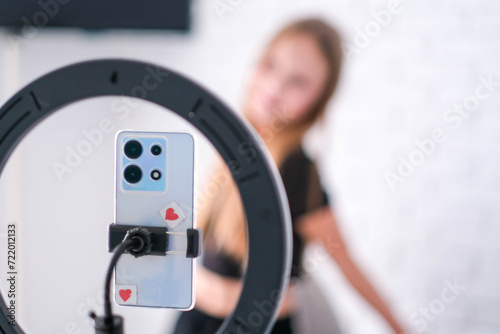 A teenage girl poses in front of a phone on a tripod. A child blogger shoots a video blog. Focus on the ring lamp and telephone.