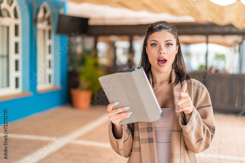Young pretty woman holding a tablet at outdoors surprised and pointing front
