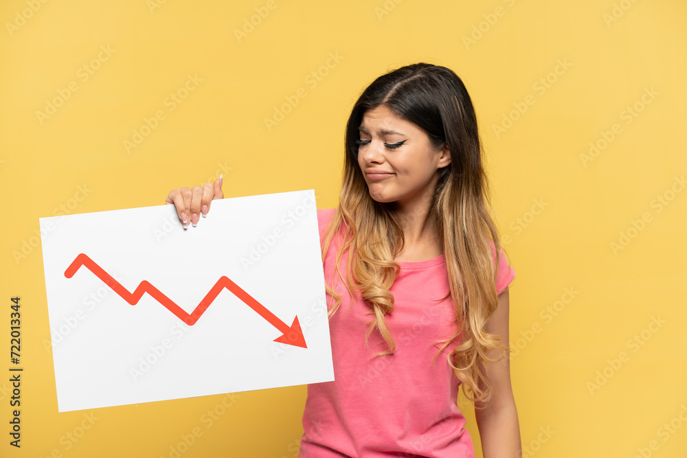 Young Russian girl isolated on yellow background holding a sign with a decreasing statistics arrow symbol with sad expression Young Russian girl isolated on yellow background holding a sign with a decreasing statistics arrow symbol with sad expression