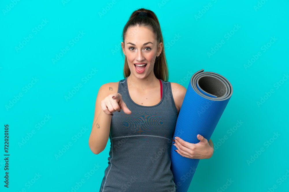 Young sport caucasian woman going to yoga classes while holding a mat isolated on blue background surprised and pointing front