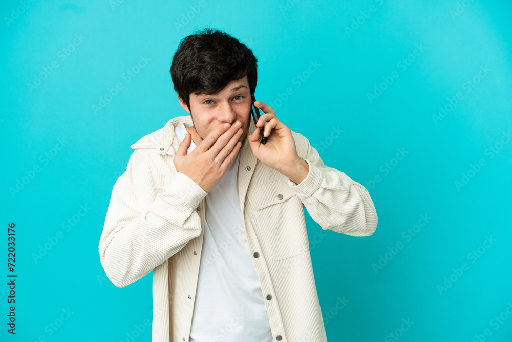 Young Russian man using mobile phone isolated on blue background happy and smiling covering mouth with hand Young Russian man using mobile phone isolated on blue background happy and smiling covering mouth with hand
