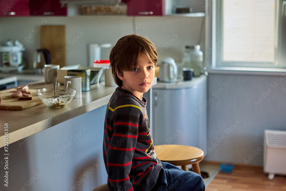 Sad, unhappy male child is bored in kitchen sitting on chair in front of furniture island. Sad, unhappy male child is bored in kitchen sitting on chair in front of furniture island.