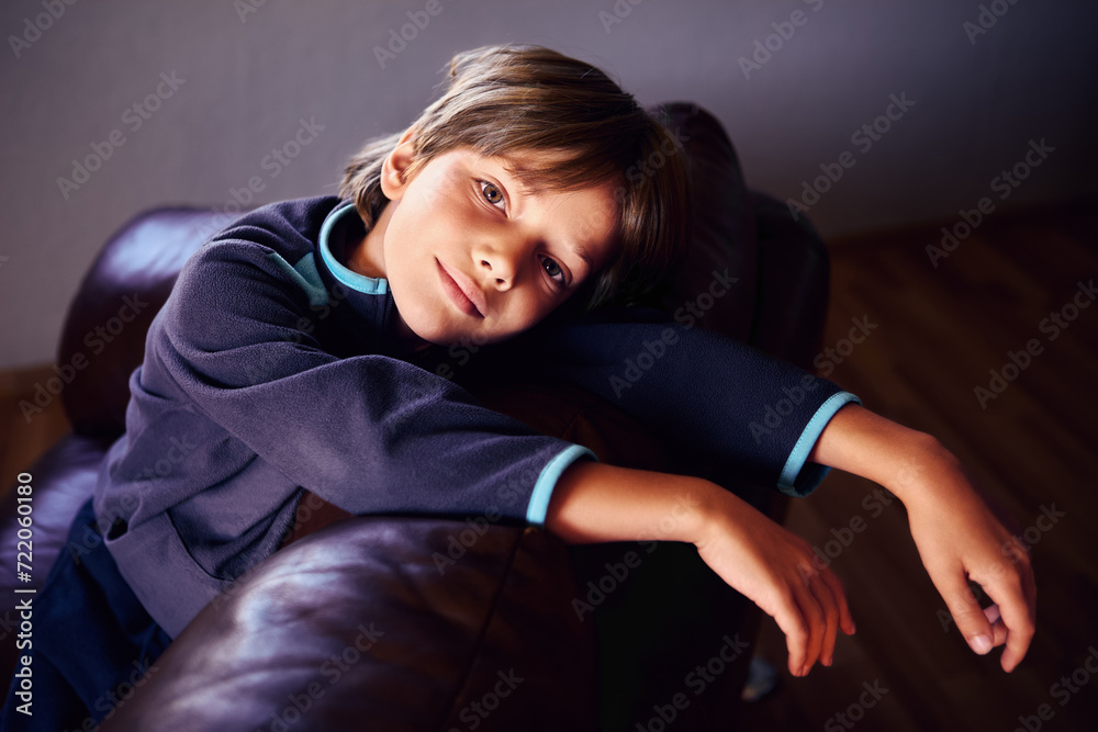 Obraz premium Sad male child is deprived of friends and gadgets as punishment for pranks, he is bored in dimly lit room alone, sitting on brown leather sofa.