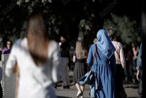 A Muslim woman wearing a blue headscarf and a blue dress walks down the street. View from the back without face