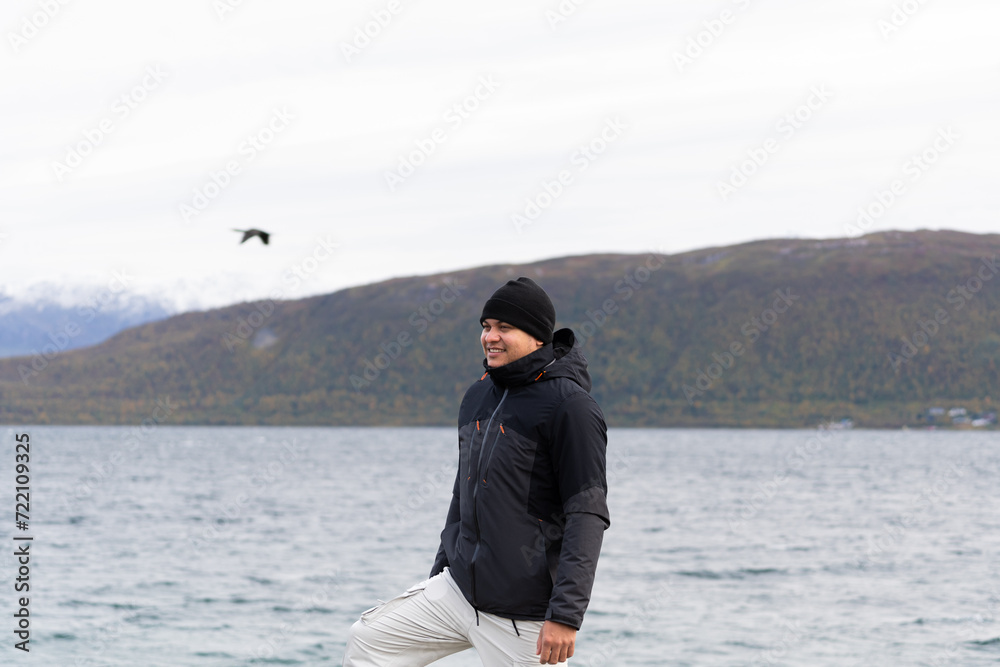 Fototapeta premium hombre latino con ropa de invierno con fondo desenfocado de fiordo noruego y ave volando