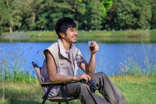 Young Asian male man sitting on chair, drinking coffee, feeling relax and raising hands, during camping beside lake and mountain.