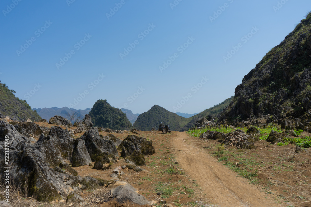 A motorcyclist riding on a remote dirt road amidst rocky terrain and rolling hills under a clear blue sky.