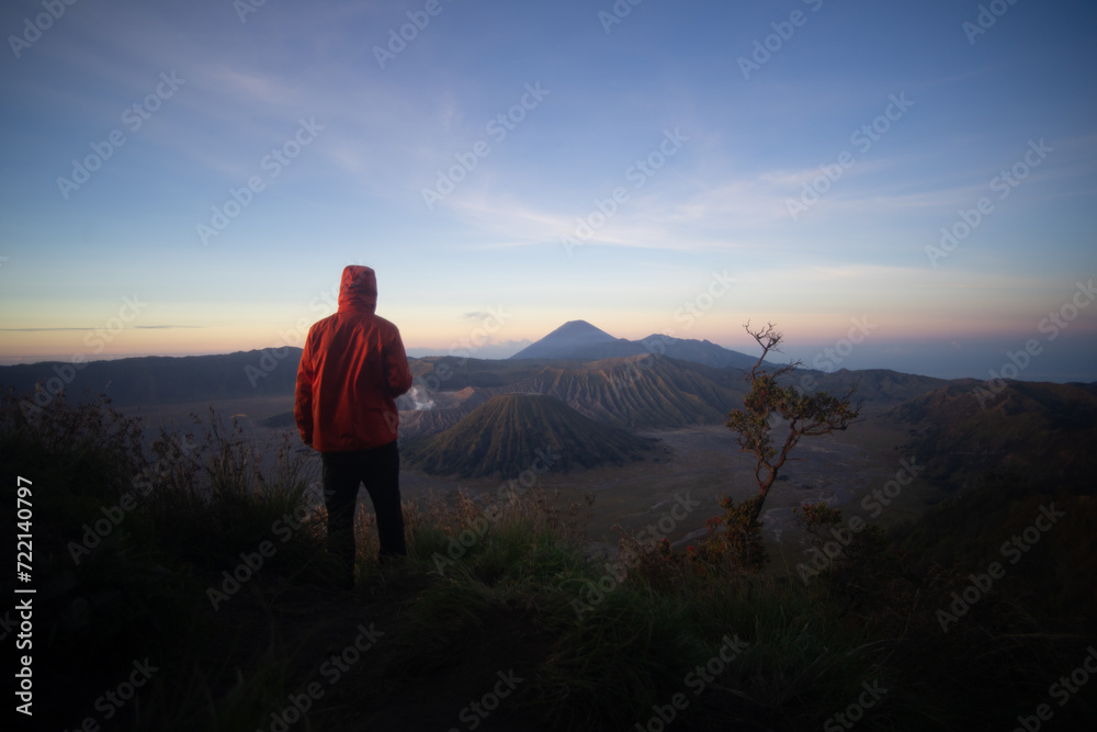 A person in a red jacket standing in front of a scenic mountain view at sunrise.