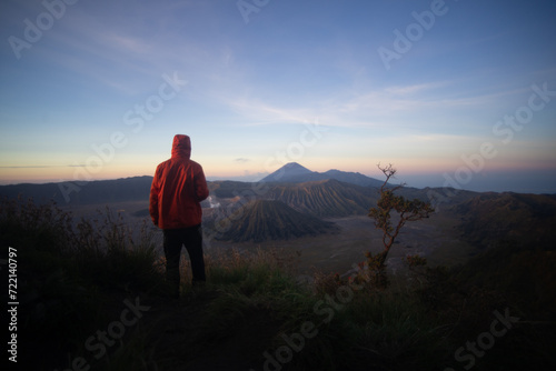 A person in a red jacket standing in front of a scenic mountain view at sunrise.