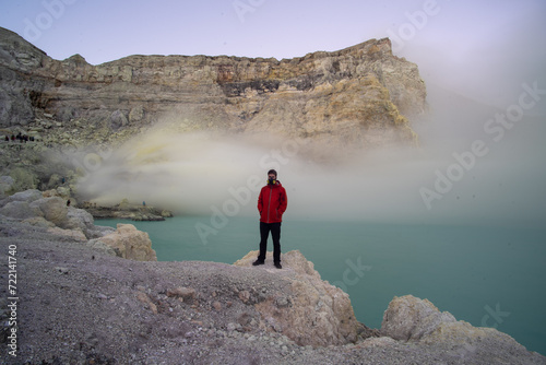 A person in a red jacket standing on the edge of a misty lake with rocky cliffs in the background.