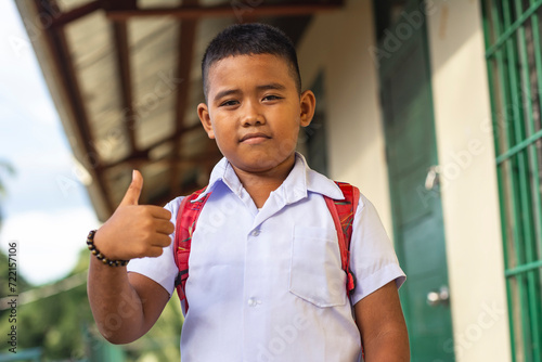 Confident young boy student with backpack outside classroom showing thumbs up, ready for school day. photo