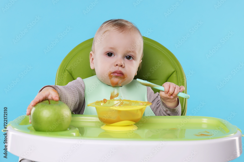 Cute little baby eating healthy food in high chair on light blue background Cute little baby eating healthy food in high chair on light blue background