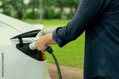 Man recharge EV electric vehicle's battery from EV charging station in outdoor green city park scenic. Eco friendly urban transport and commute with eco friendly EV car travel. Exalt