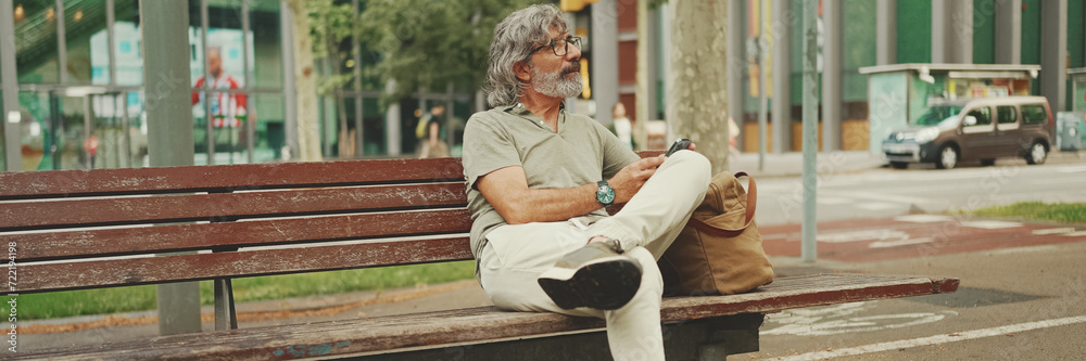 Fototapeta premium Panorama of positive middle-aged man with gray hair and beard wearing casual clothes sits on bench and uses mobile phone. Mature gentleman in eyeglasses writes message on smartphone