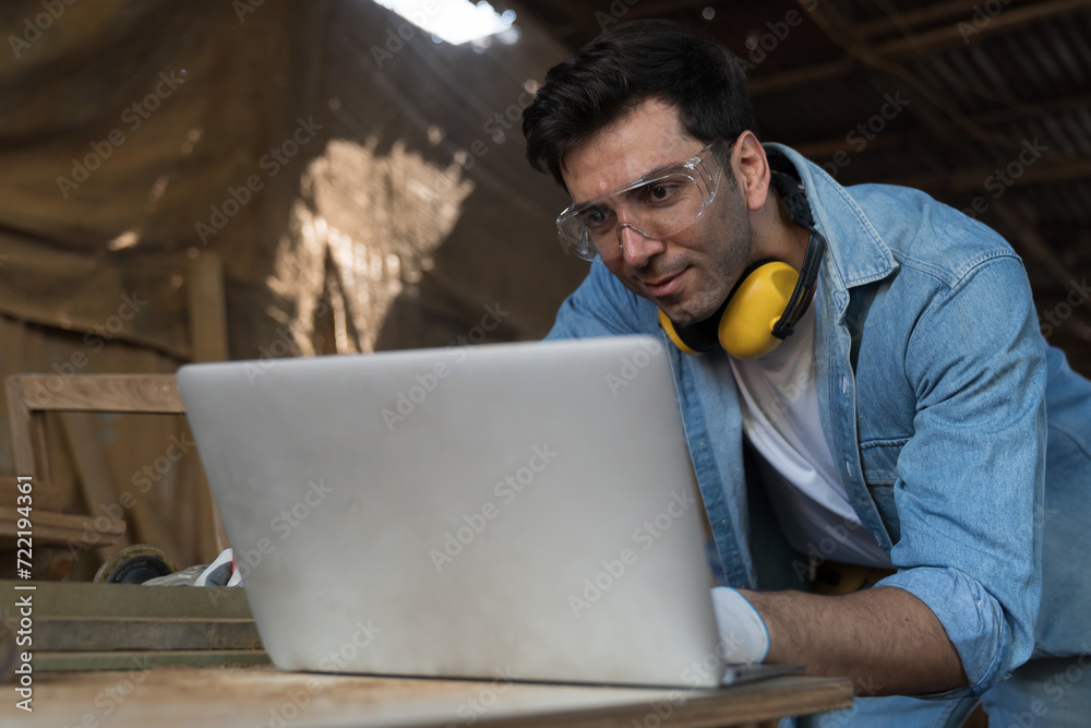Male carpenter using laptop computer at woodwork workshop. Male joiner working with laptop computer furniture workshop. Start up and small business concept Male carpenter using laptop computer at woodwork workshop. Male joiner working with laptop computer furniture workshop. Start up and small business concept