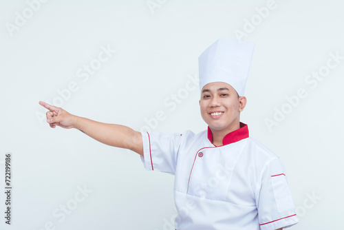 Happy Asian male chef in uniform pointing to the side, isolated on a white background, conveying a friendly gesture.
