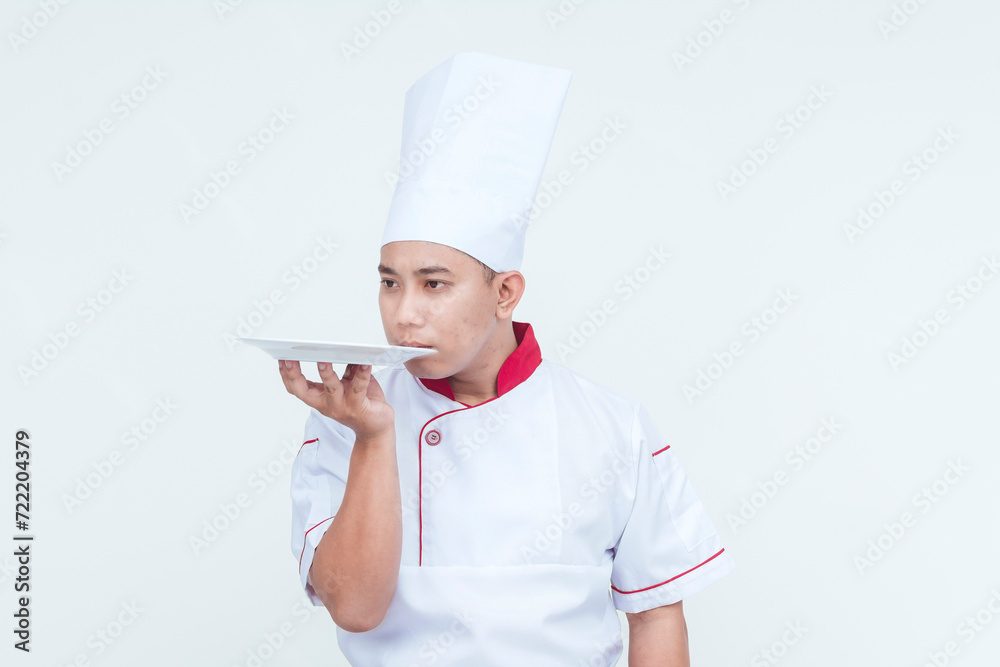 Focused Asian chef in professional attire smelling an empty plate, isolated against a white background with copy space. Focused Asian chef in professional attire smelling an empty plate, isolated against a white background with copy space.