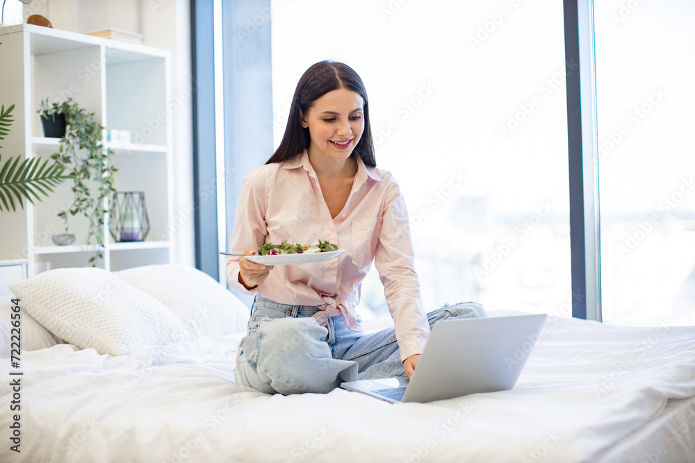 Relaxed caucasian woman in casual wear sitting on comfy bed with wireless laptop, eating healthy salad from fresh vegetables for breakfast on background of light modern cozy bedroom.