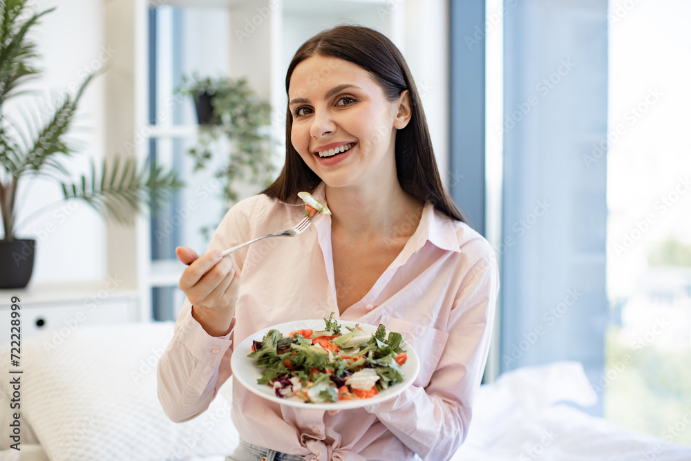 Close up portrait of caucasian woman in casual wear sitting on comfy bed eating healthy salad. Brunette female enjoying breakfast from fresh vegetables on background of light modern cozy bedroom.