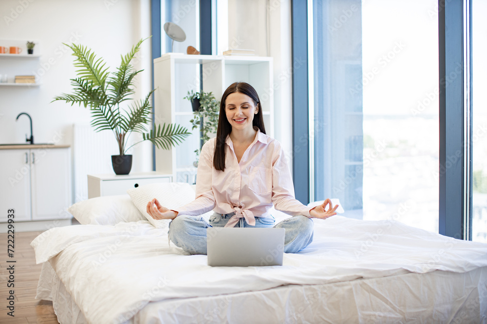 Peaceful lady sitting with closed eyes on bed on lotus position during online video class using modern laptop. Caucasian woman enjoying practicing yoga during break with modern laptop at home.