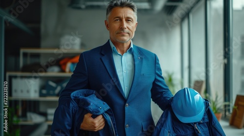 Downshifting office workers into blue-collar workers. A middle-aged man in a classic blue suit holds a blue work jacket and helmet in his hands and is preparing to change clothes. photo