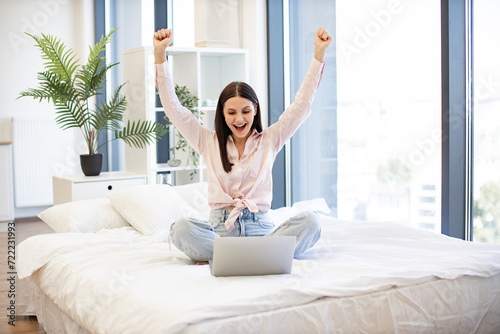 Happy young woman in casual wear raising hands up while using laptop for ordering new cosmetics during sale season. Online shopping with discount at home. E-commerce and technology concept.