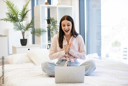 Front view of smiling woman having video chat with friends on laptop while sitting on comfortable bed. Charming female enjoying communication at distance using modern technologies while staying home.