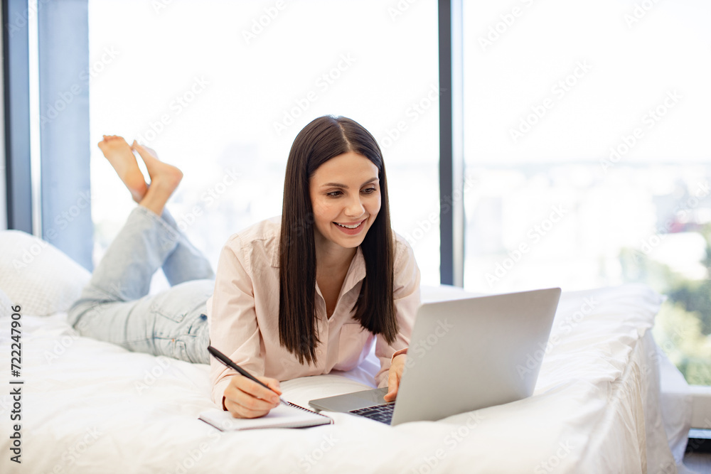 Beautiful young woman relaxing on cozy bed at home using modern laptop while writing in notebook with pen. Caucasian brunette student enjoying studying at home on weekend. Beautiful young woman relaxing on cozy bed at home using modern laptop while writing in notebook with pen. Caucasian brunette student enjoying studying at home on weekend.