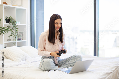 Happy young woman holding wireless joystick while playing video games on modern laptop. Happy lady sitting on comfy bed and enjoying favorite computer game on weekend.