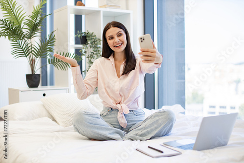 Beautiful lady sitting on comfy bed and enjoying online conversation with relatives or friends on weekend. Caucasian young woman holding wireless smartphone having a video call.